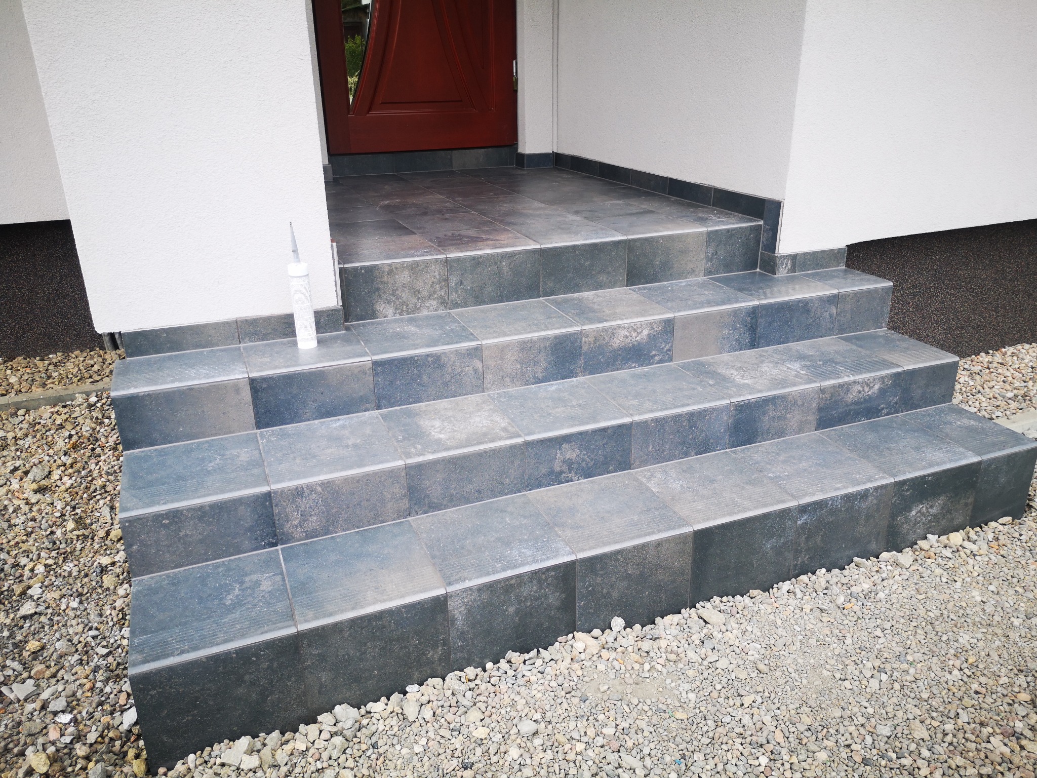 Wykończone schody wejściowe do domu, wyłożone ciemnymi płytkami gresowymi, z widoczną tubą silikonu na górnym stopniu i żwirowym podłożem.