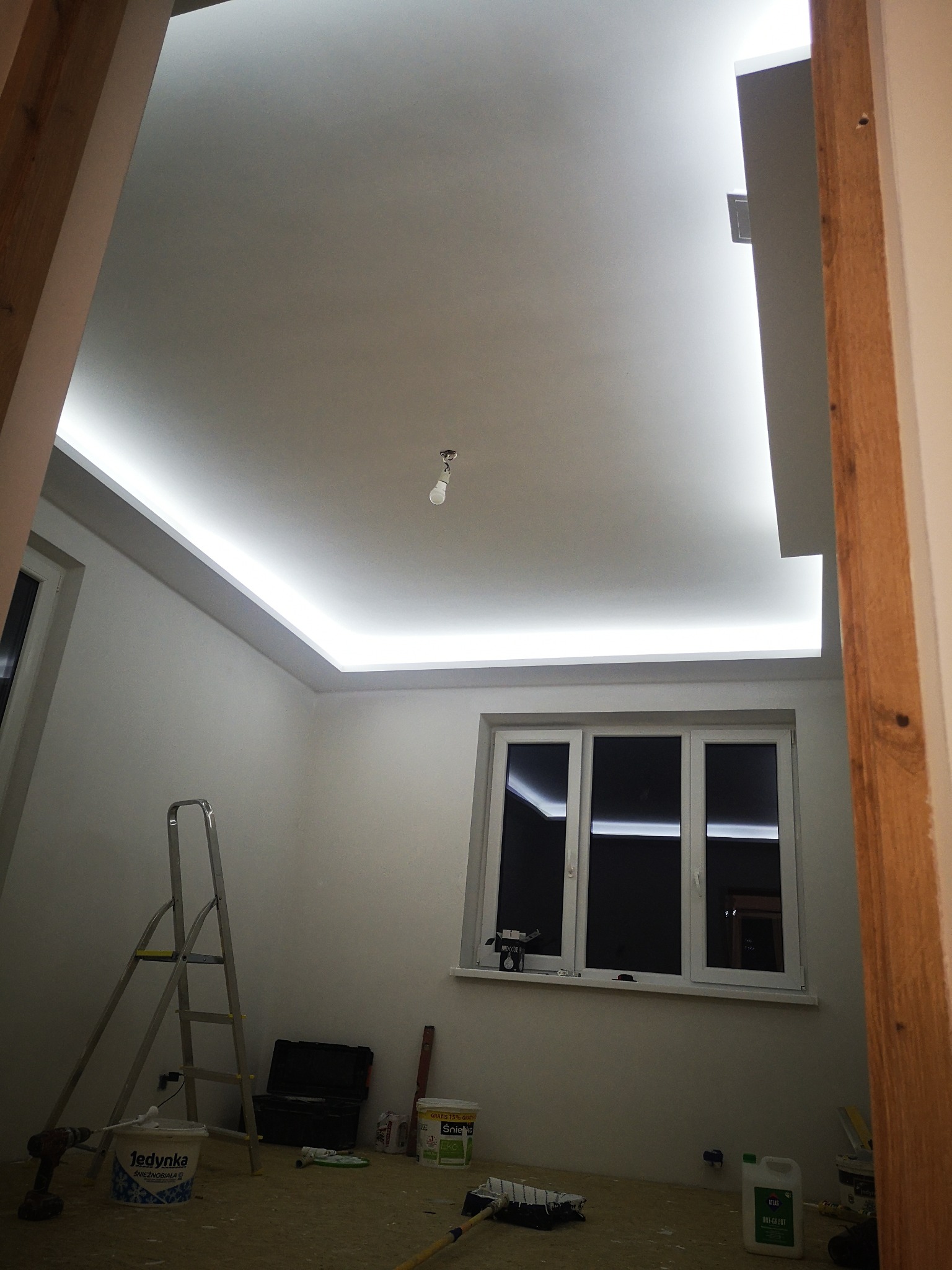 Pokój w trakcie remontu z podwieszanym sufitem z oświetleniem LED, drabina, narzędzia malarskie i wiadro z farbą 'Jedynka' na podłodze.