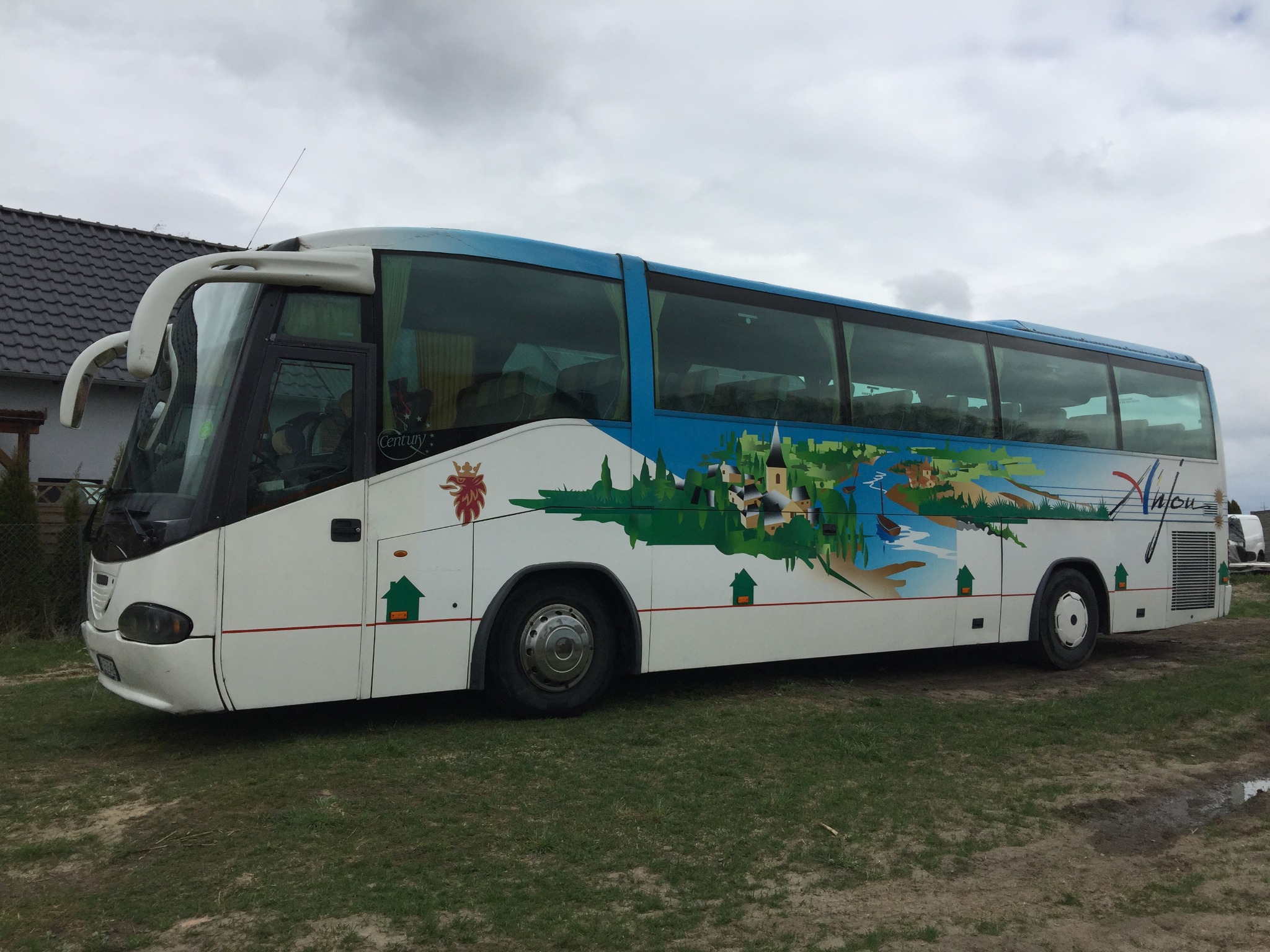 Autokar Scania z grafiką krajobrazu i logo firmy na boku, stojący na trawiastym terenie.