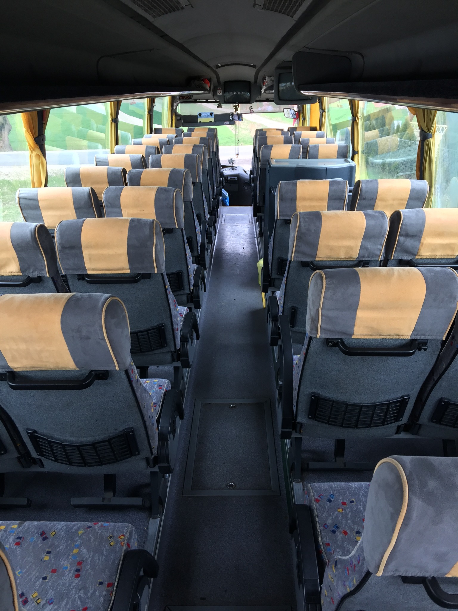 Wnętrze autobusu Scania z rzędami szarych foteli z żółtymi zagłówkami, widok z przodu na puste siedzenia i okna z zasłonkami.