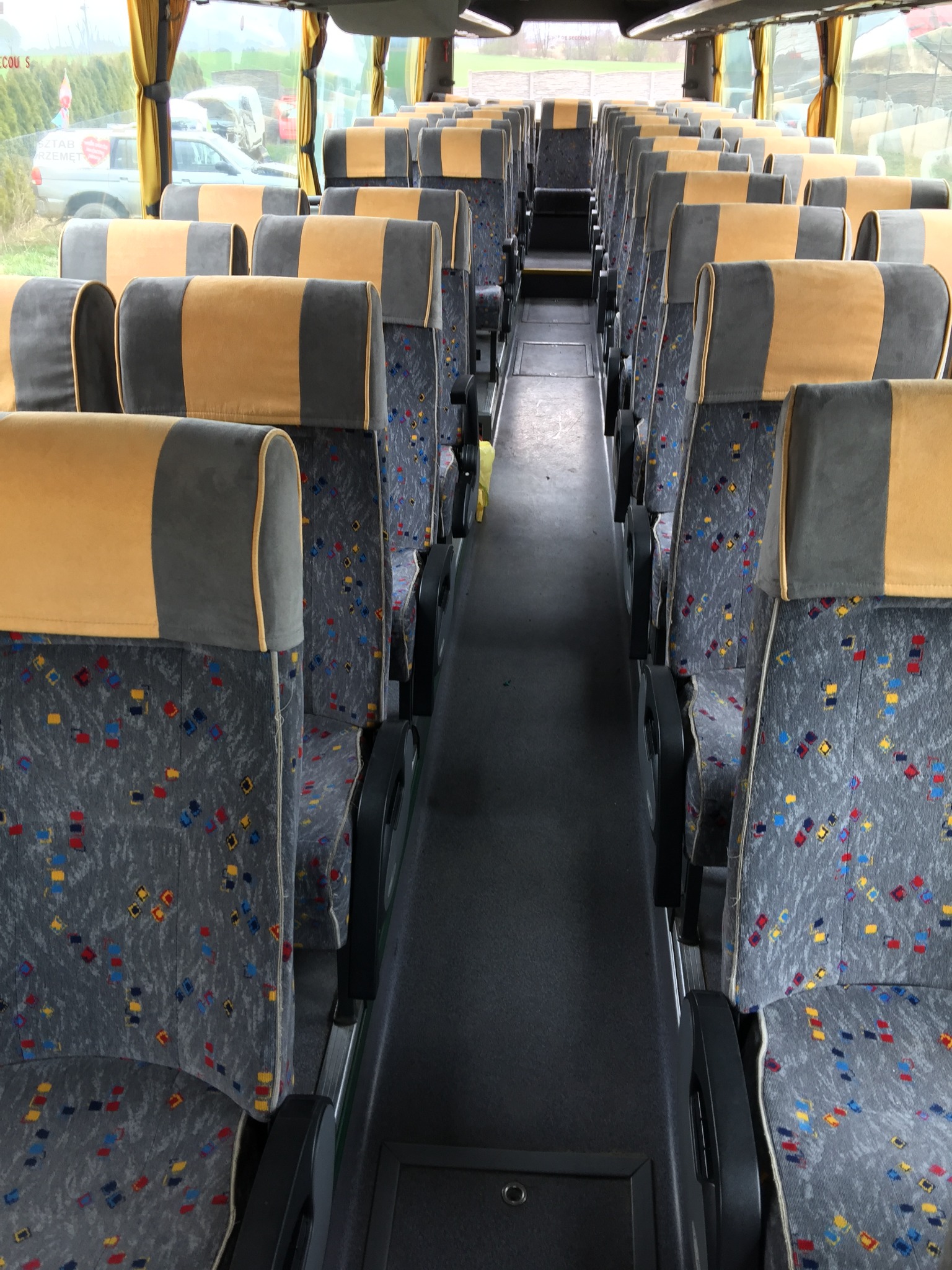 Wnętrze autobusu z rzędami szarych siedzeń z kolorowymi wzorami i żółto-szarymi zagłówkami, widok wzdłuż korytarza.