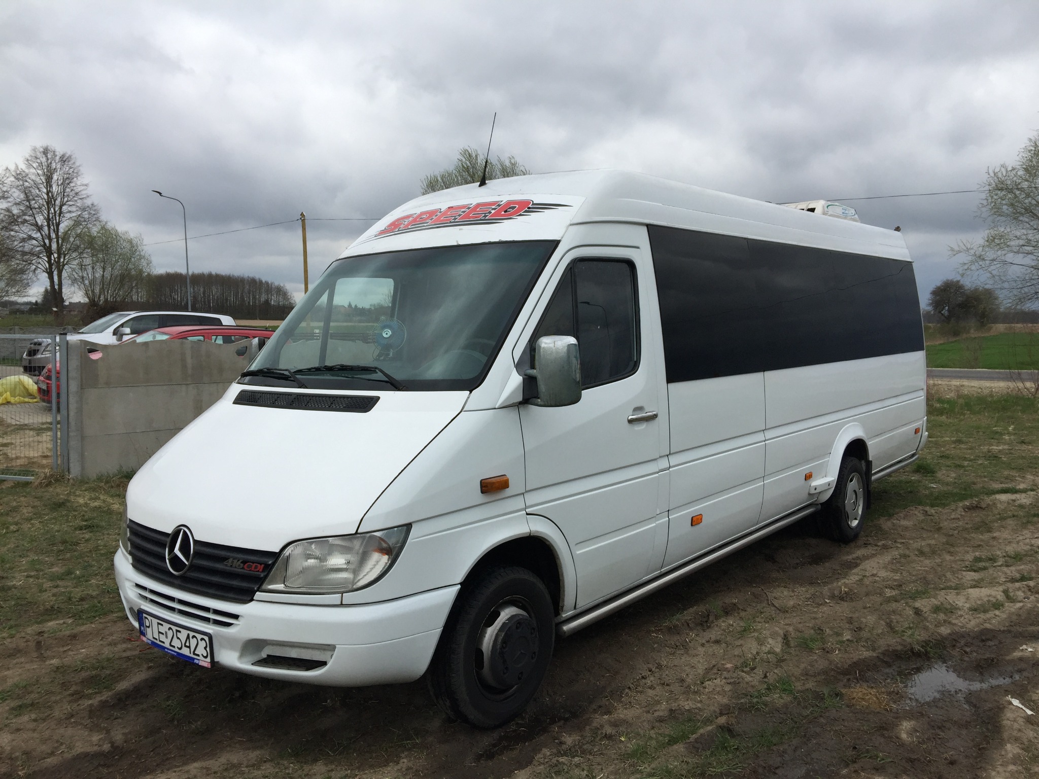 Biały bus Mercedes-Benz Sprinter z czarnymi szybami i czerwonym napisem 'SPEED' na dachu, stojący na błotnistym terenie pod pochmurnym niebem.