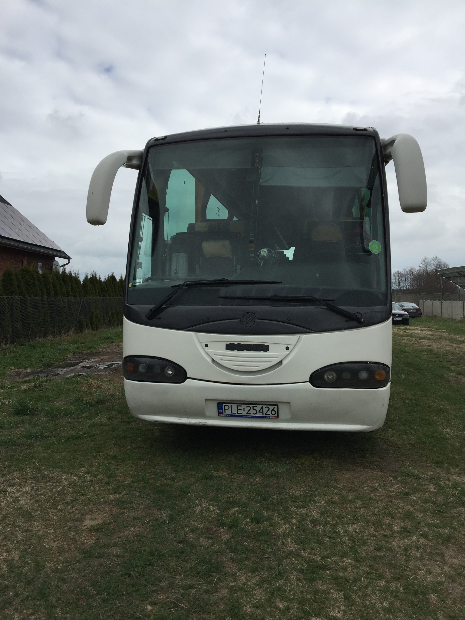 Biały autokar marki SCANIA z numerem rejestracyjnym PLE 25426, zaparkowany na trawie przed budynkiem z panelami słonecznymi.