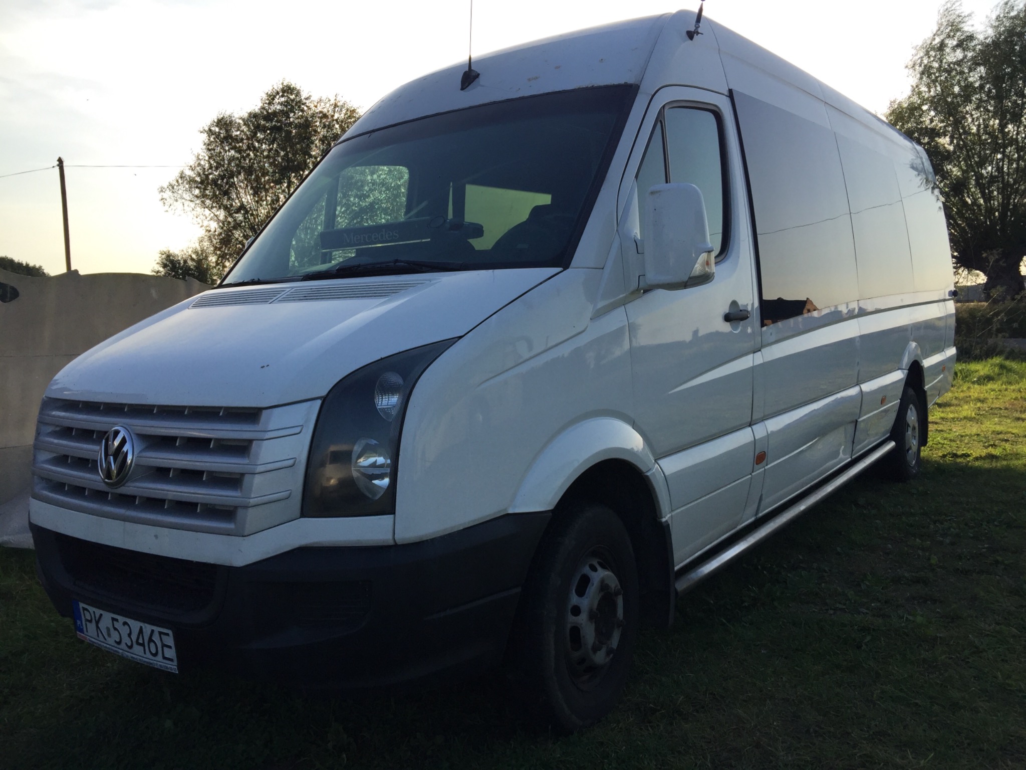 Biały bus Volkswagen Crafter z przyciemnianymi szybami, zaparkowany na trawie, z widoczną tablicą rejestracyjną PK 5346E i naklejką Mercedes na przedniej szybie.