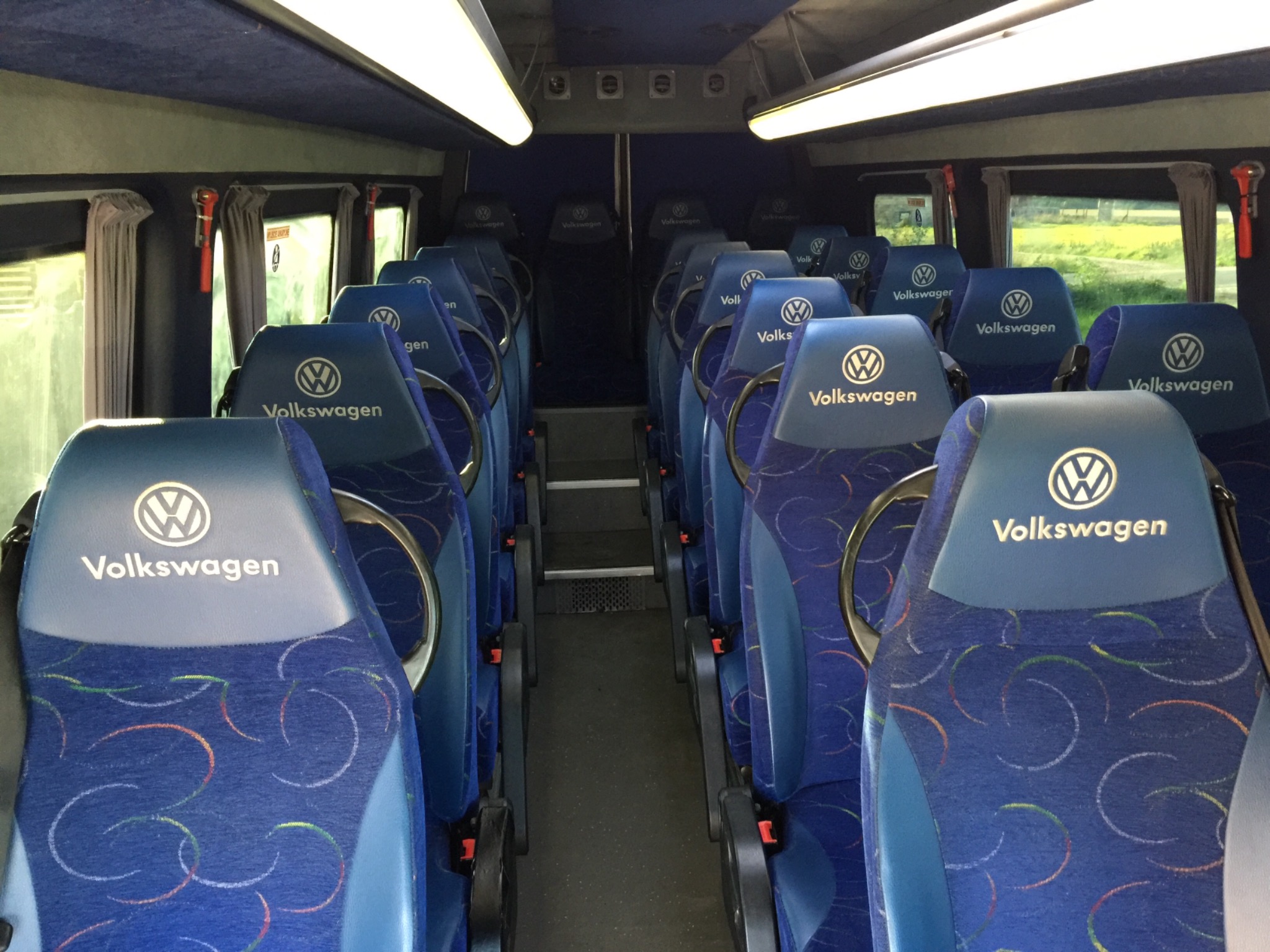 Wnętrze autobusu Volkswagen z rzędami niebieskich foteli z logo marki, widok wzdłuż przejścia.