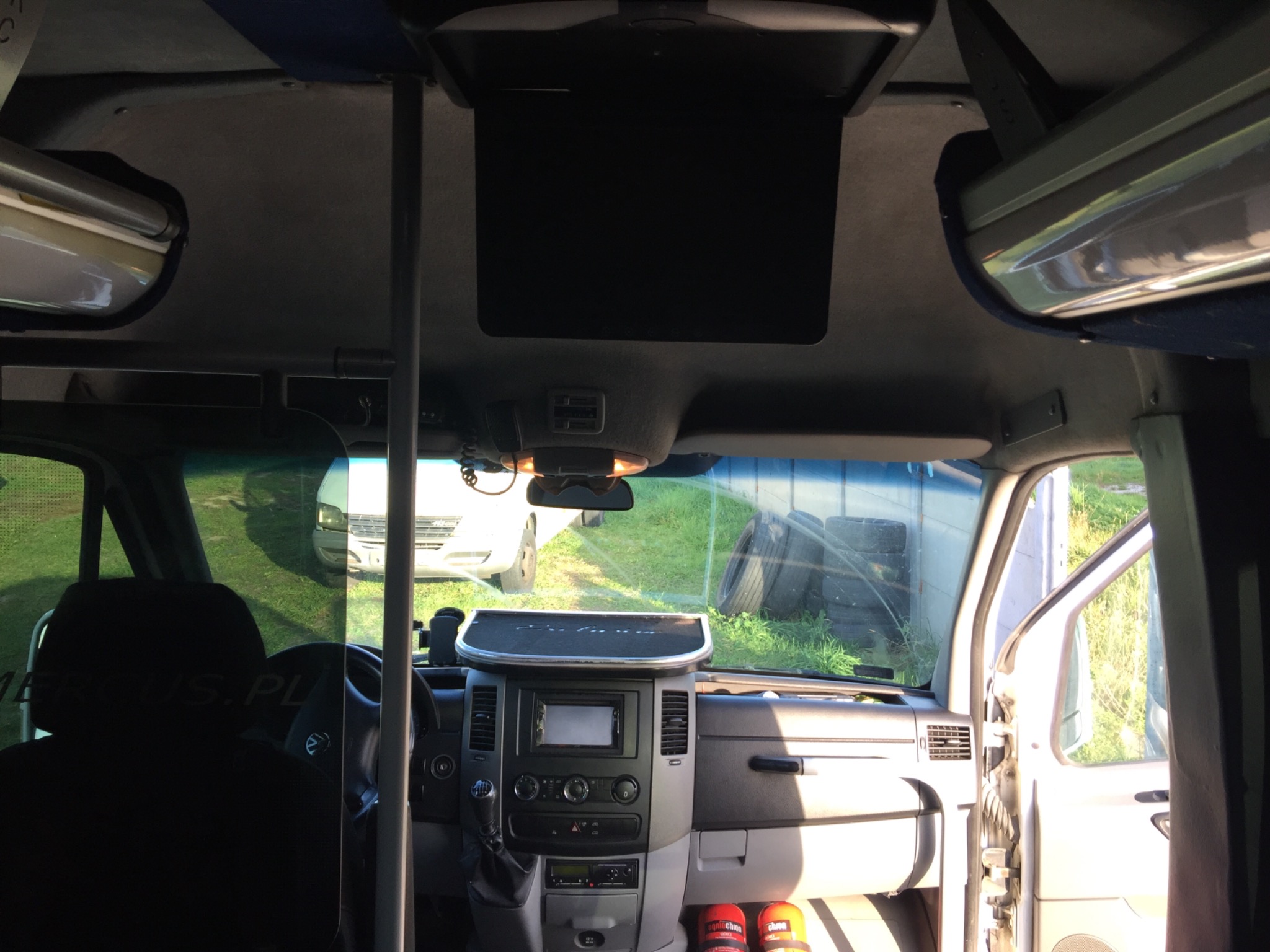 Wnętrze busa z ekranem sufitowym, widok z tyłu kabiny, deska rozdzielcza z radiem i klimatyzacją, widoczne opony na zewnątrz przez przednią szybę.