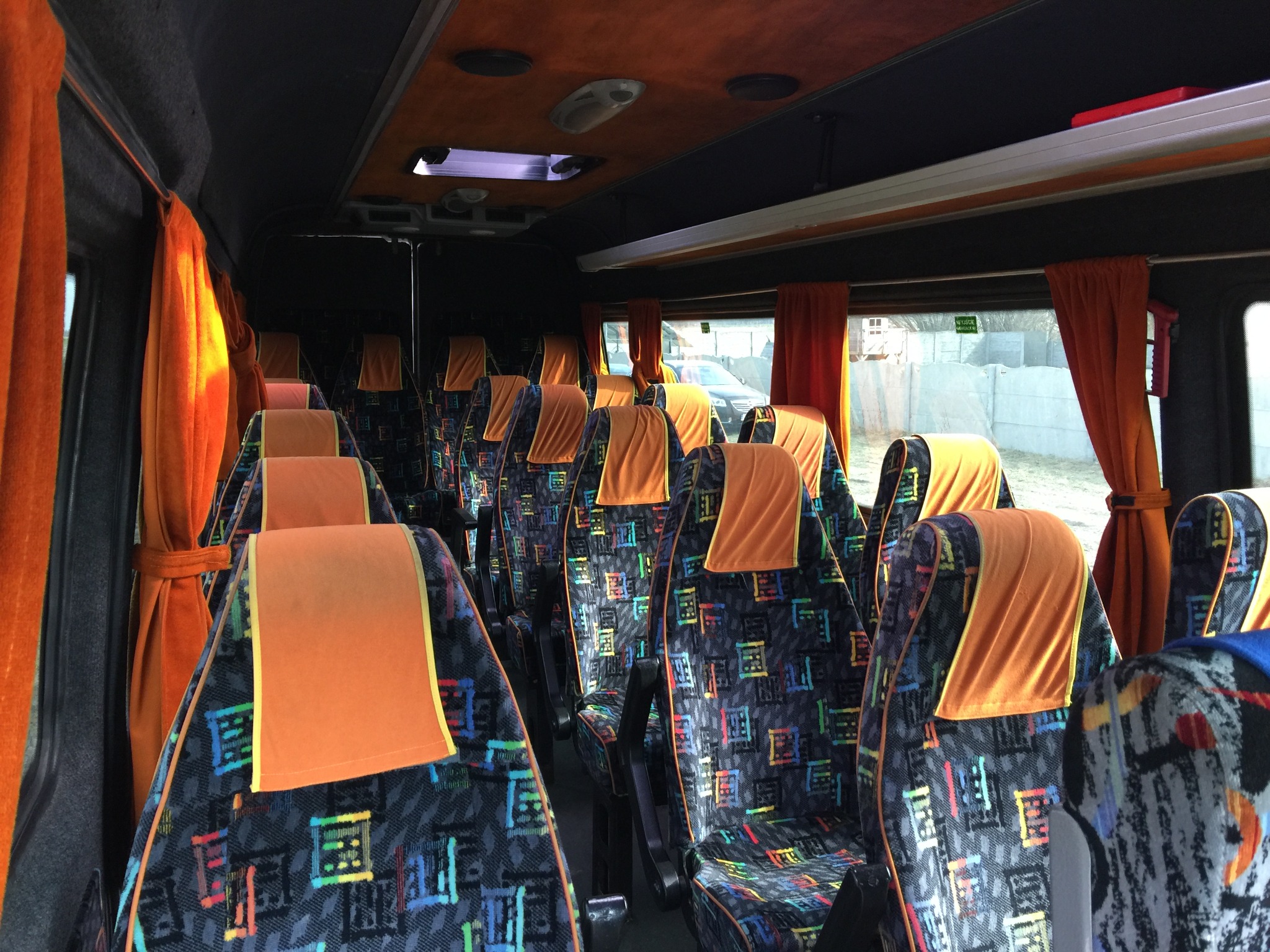 Wnętrze autobusu z ciemną tapicerką w kolorowe wzory, pomarańczowe zasłony i zagłówki, widok z przodu na puste siedzenia.