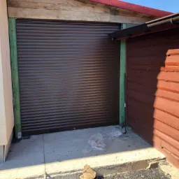 Zamknięta, brązowa roleta garażowa widziana z zewnątrz, z elementami konstrukcyjnymi w kolorze zielonym i fragmentami drewnianej elewacji po prawej stronie, na betonowym podłożu leżą resztki folii...