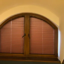 Arched window with closed, terracotta-colored pleated shades, dark wood frame, and dark sill. The wall is painted a warm yellow.