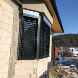 Montaż żaluzji fasadowych zewnętrznych w surowym budynku, widoczne bloczki betonowe, ocieplenie i fragment dachu, w tle las i błękitne niebo.