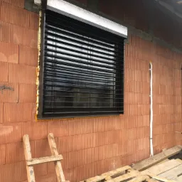 Ciemne żaluzje fasadowe zamontowane w oknie budynku w trakcie budowy, widoczna cegła ceramiczna, pianka montażowa i drewniana drabina oparta o ścianę.