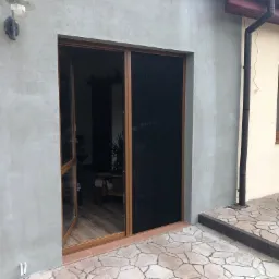 Zewnętrzne drzwi z brązową ramą i zamontowaną moskitierą przesuwną, widok z zewnątrz na szarą elewację budynku i fragment tarasu z kamienną posadzką.