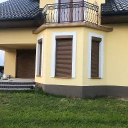 Żółty dom z brązowymi roletami zewnętrznymi, widok z trawnika, z garażem zamykanym roletą.