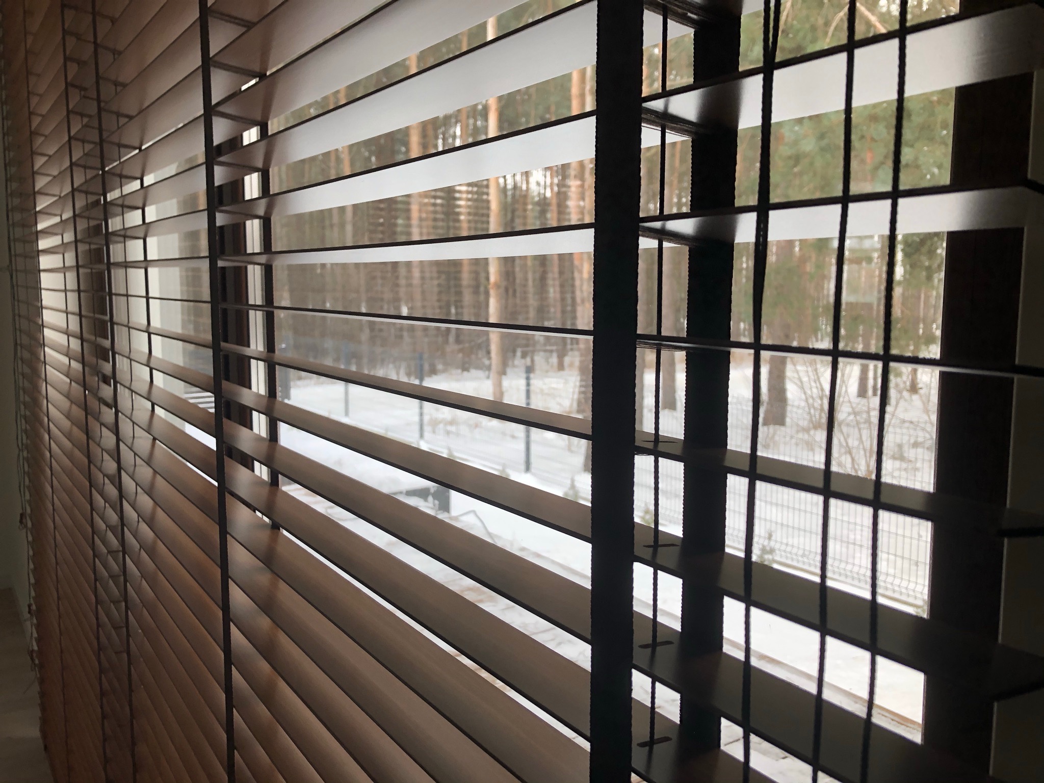 Drewniane żaluzje poziome z widokiem na zimowy krajobraz za oknem, częściowo zasłonięte, z widoczną fakturą drewna