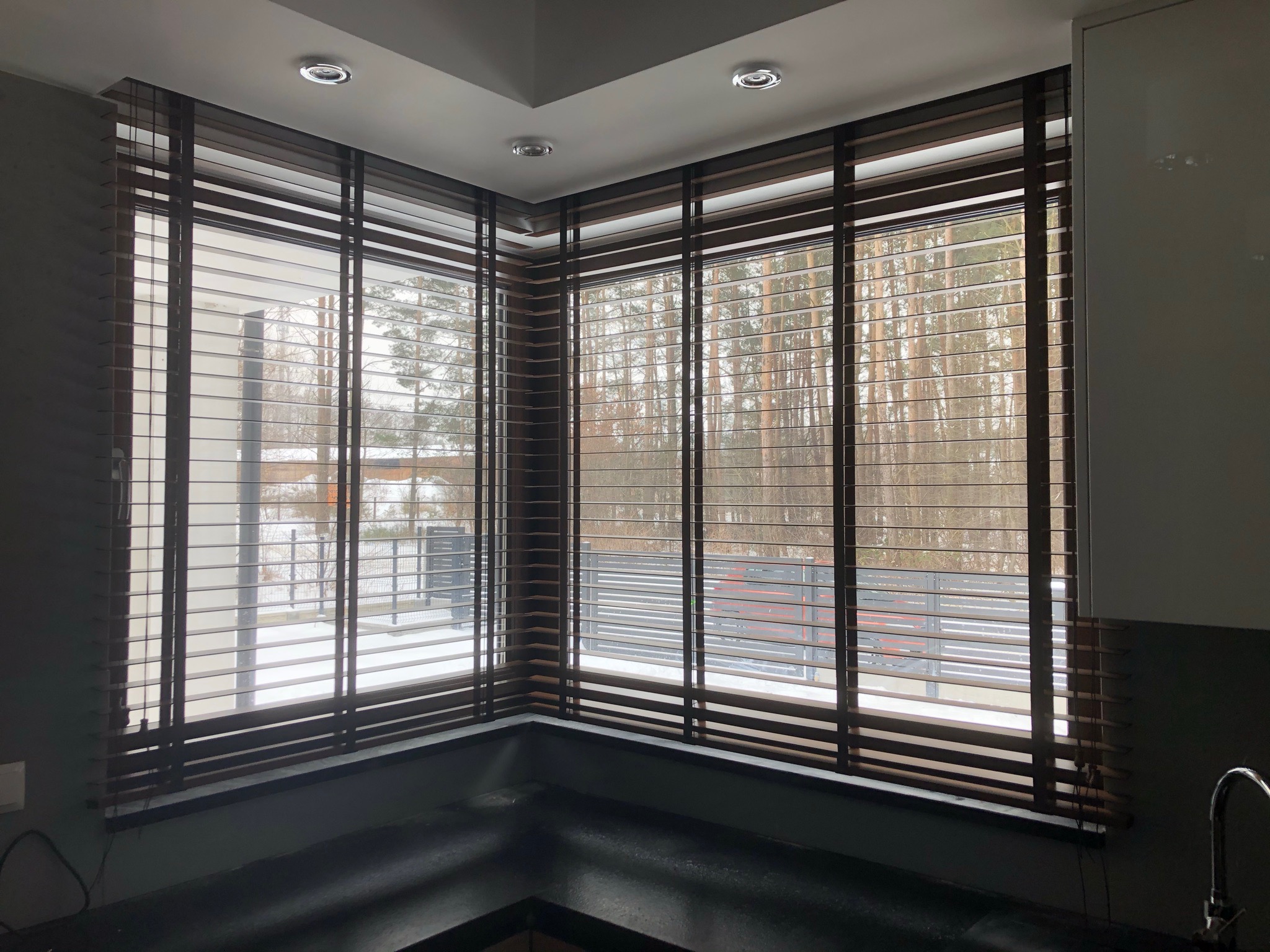 Drewniane żaluzje poziome w narożnym oknie z widokiem na zimowy krajobraz z drzewami i ogrodzeniem.