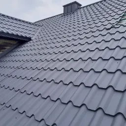 Nowy, szary dach z blachodachówki modułowej z kominem wentylacyjnym, widok z dołu na tle pochmurnego nieba.