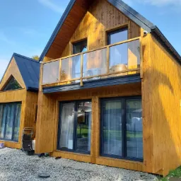 Nowoczesny, drewniany dom z balkonem zabezpieczonym szklaną balustradą i czarnym dachem, widok z zewnątrz na żwirowe podłoże.