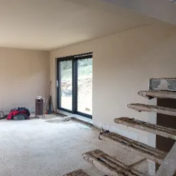 Wnętrze w trakcie tynkowania maszynowego, widoczne świeżo otynkowane ściany, surowe schody z metalowymi elementami i widok na zewnątrz przez duże okno przesuwne.