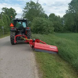 Czerwony traktor z kosiarką rotacyjną przycina trawę na poboczu drogi w Siedliszowicach, widok z tyłu, w tle drzewa i pochmurne niebo.