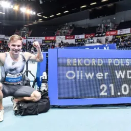 Młody sportowiec, Oliwer Wdowik, świętuje ustanowienie rekordu Polski U20 w biegu, siedząc obok tablicy wyników z czasem 21.09, ubrany w strój sportowy z numerem 197, na tle trybun hali sportowej.