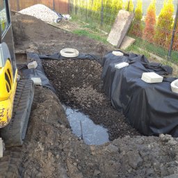 Krystian Mik - Żółta mini koparka obok wykopu z czarną folią, wypełnionego gruzem i częściowo wodą, z betonowymi kręgami i bloczkami na wierzchu, w tle ogrodzenie z siatki i tuje.