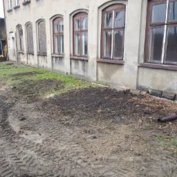 Przygotowany teren pod budynkiem z widocznymi śladami opon i małą koparką w tle, sugerujący prace ziemne i niwelację terenu.