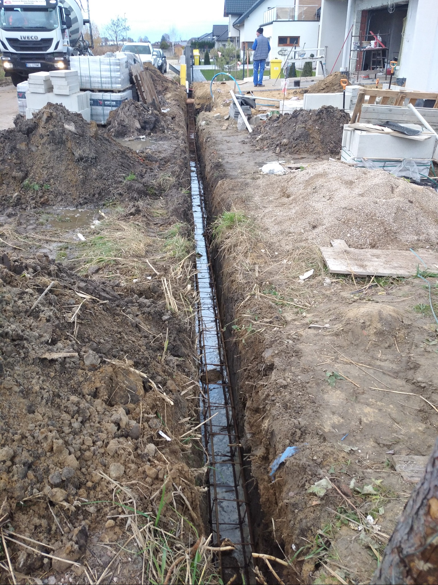 Długi, wąski wykop wypełniony wodą i zbrojeniem stalowym, przygotowany pod fundament. Na pierwszym planie sterty ziemi, w tle betoniarka i materiały budowlane na posesji.
