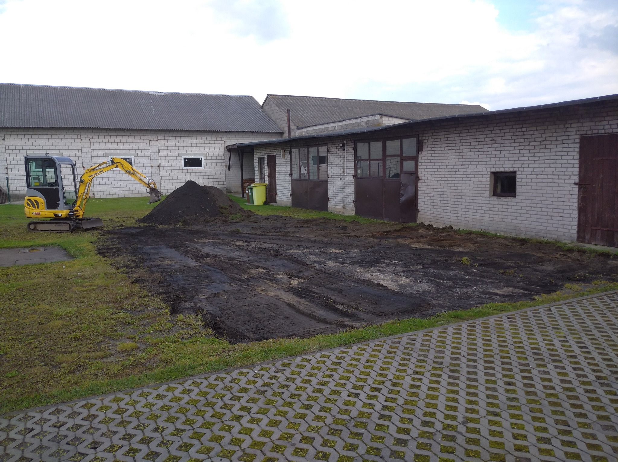 Żółta mini koparka przy stercie ziemi na placu budowy, obok budynki gospodarcze z białej cegły