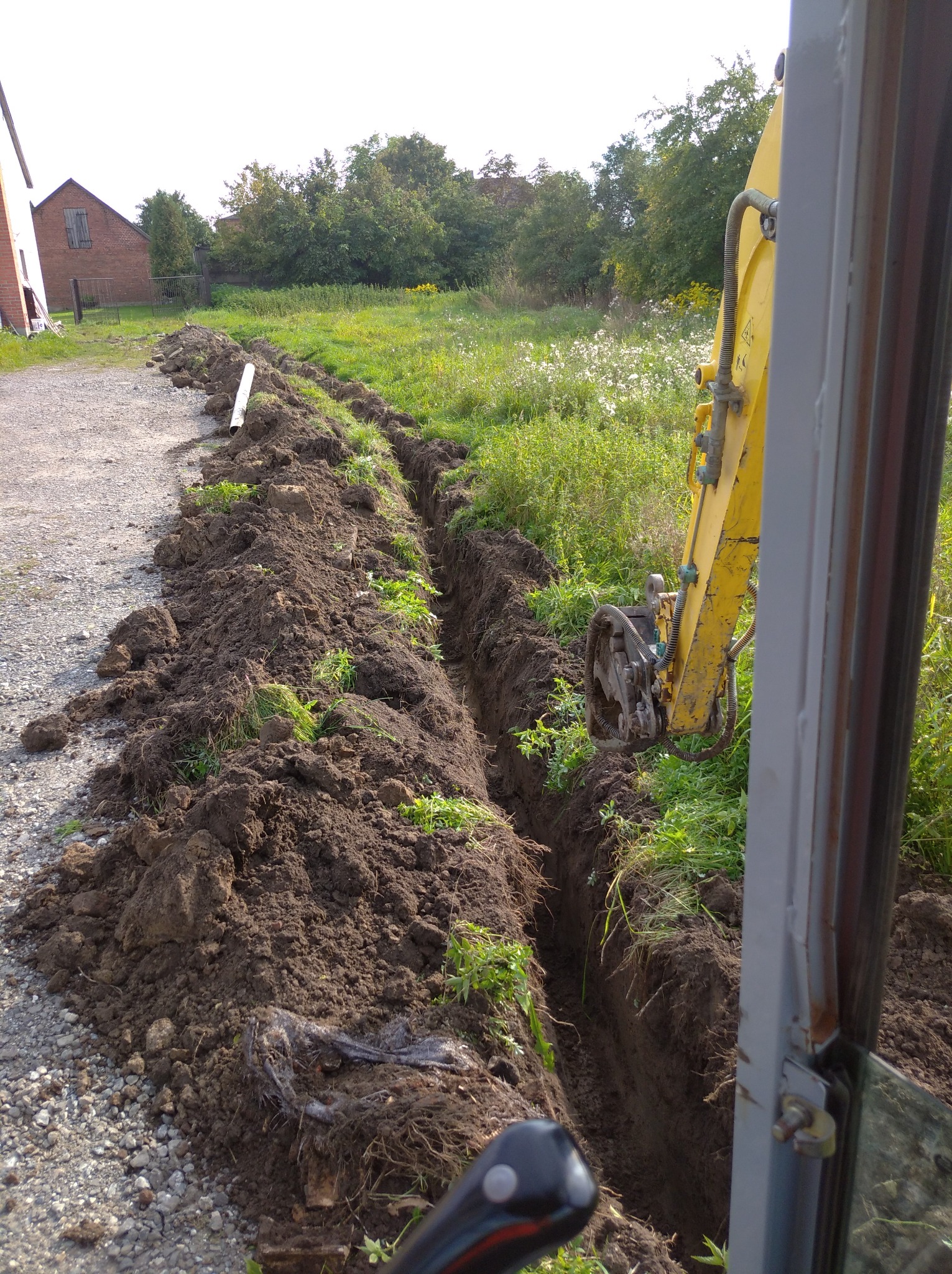 Wąski wykop w ziemi, z żółtą maszyną budowlaną częściowo widoczną po prawej stronie, widok z kabiny operatora, w tle zielona trawa i drzewa.