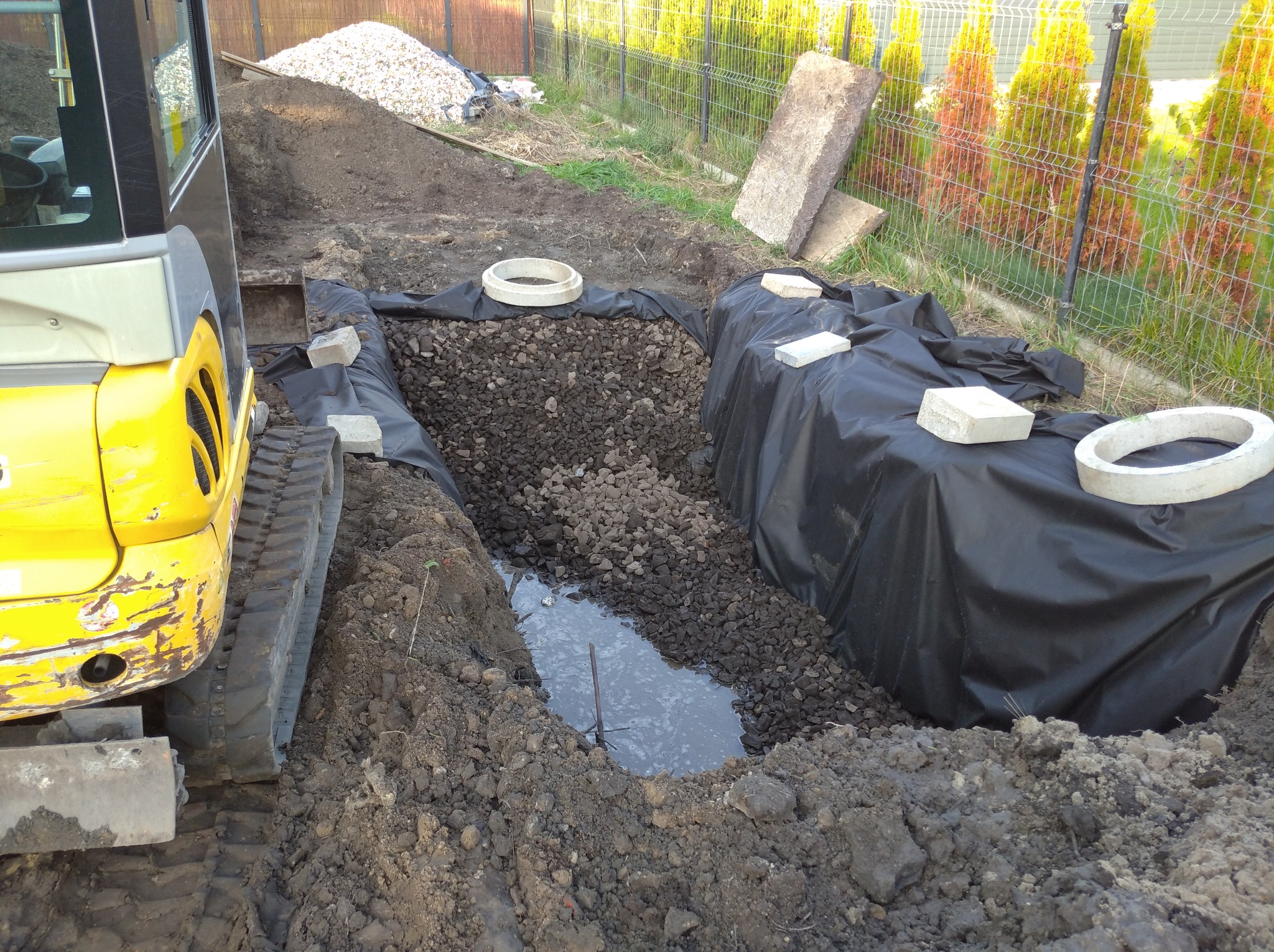 Żółta mini koparka obok wykopu z czarną folią, wypełnionego gruzem i częściowo wodą, z betonowymi kręgami i bloczkami na wierzchu, w tle ogrodzenie z siatki i tuje.