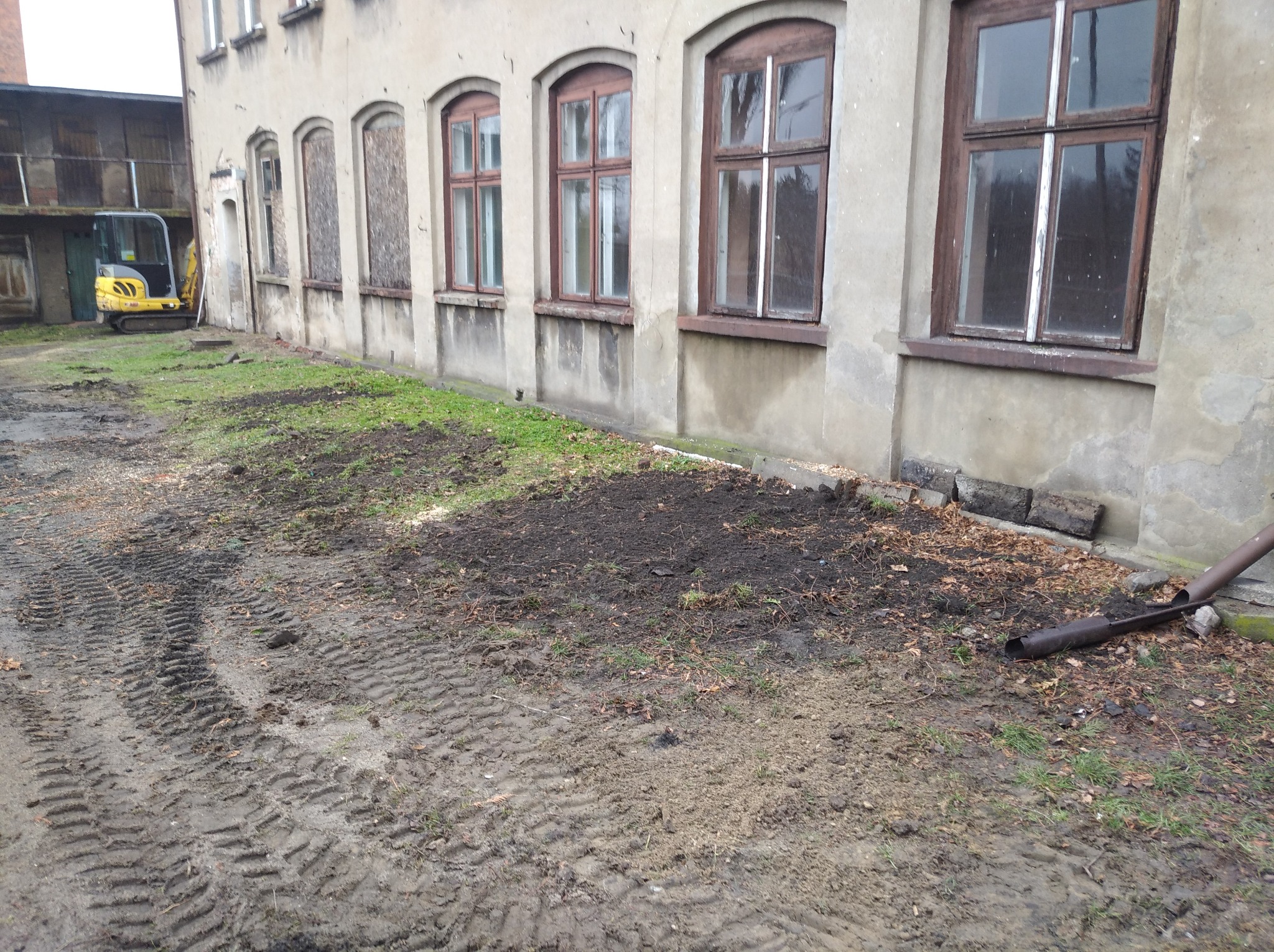 Przygotowany teren pod budynkiem z widocznymi śladami opon i małą koparką w tle, sugerujący prace ziemne i niwelację terenu.
