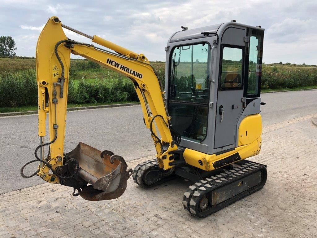 Żółto-szara minikoparka New Holland na gąsienicach, zaparkowana na poboczu asfaltowej drogi, z zielonym polem i drzewami w tle.