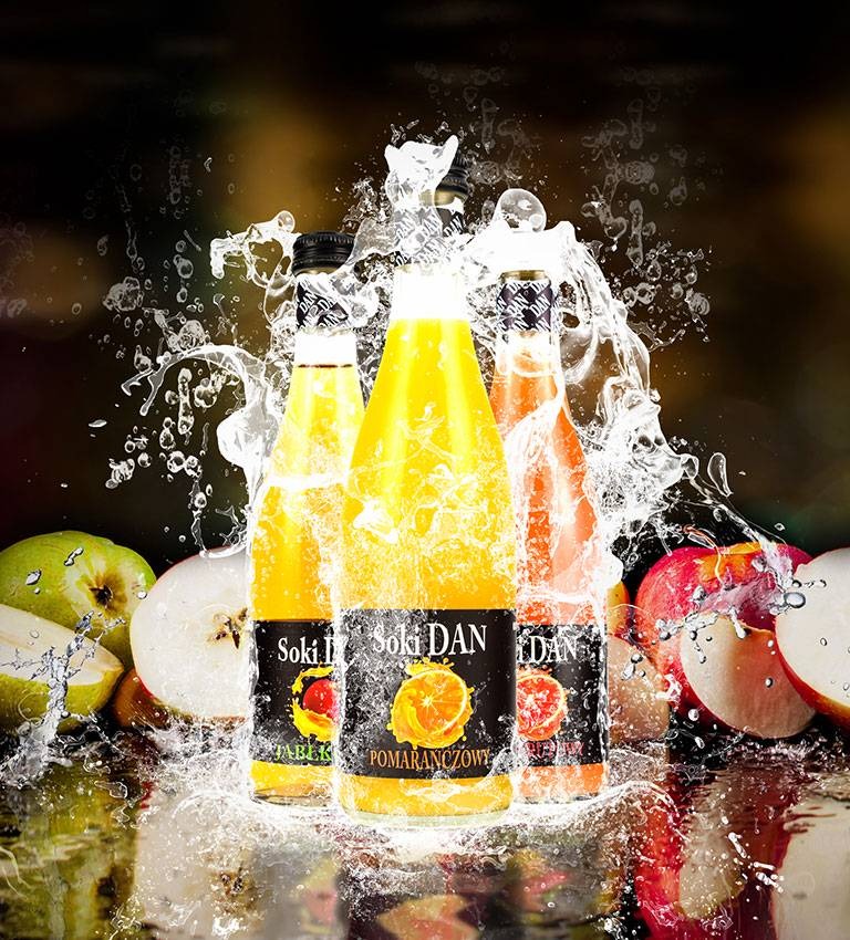 Trzy szklane butelki z sokiem owocowym (jabłkowy, pomarańczowy, grejpfrutowy) marki Soki DAN, otoczone rozpryskującą się wodą i połówkami jabłek i pomarańczy na błyszczącej powierzchni.