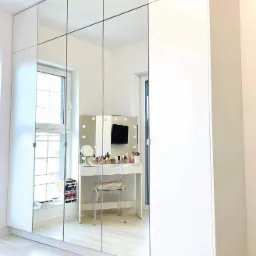 Biała szafa wnękowa z lustrzanymi frontami, odbijająca toaletkę z oświetleniem i przezroczyste krzesło, na jasnej podłodze.