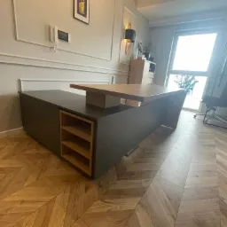 Nowoczesne biurko z ciemnoszarym korpusem i blatem z jasnego drewna, z otwartymi p&oacute;łkami, ustawione na parkiecie w jodełkę, w tle ściana z dekoracyjnymi listwami.