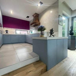 TMP Meble - Nowoczesna kuchnia z szarymi szafkami, fioletowymi górnymi szafkami, drewnianym okapem i wyspą z dekoracjami, widoczna z poziomu podłogi z desek.