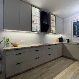 Nowoczesna kuchnia z szarymi szafkami, czarnymi uchwytami, przeszklonymi witrynami z podświetleniem LED, drewnianym blatem i drewnopodobną podłogą.