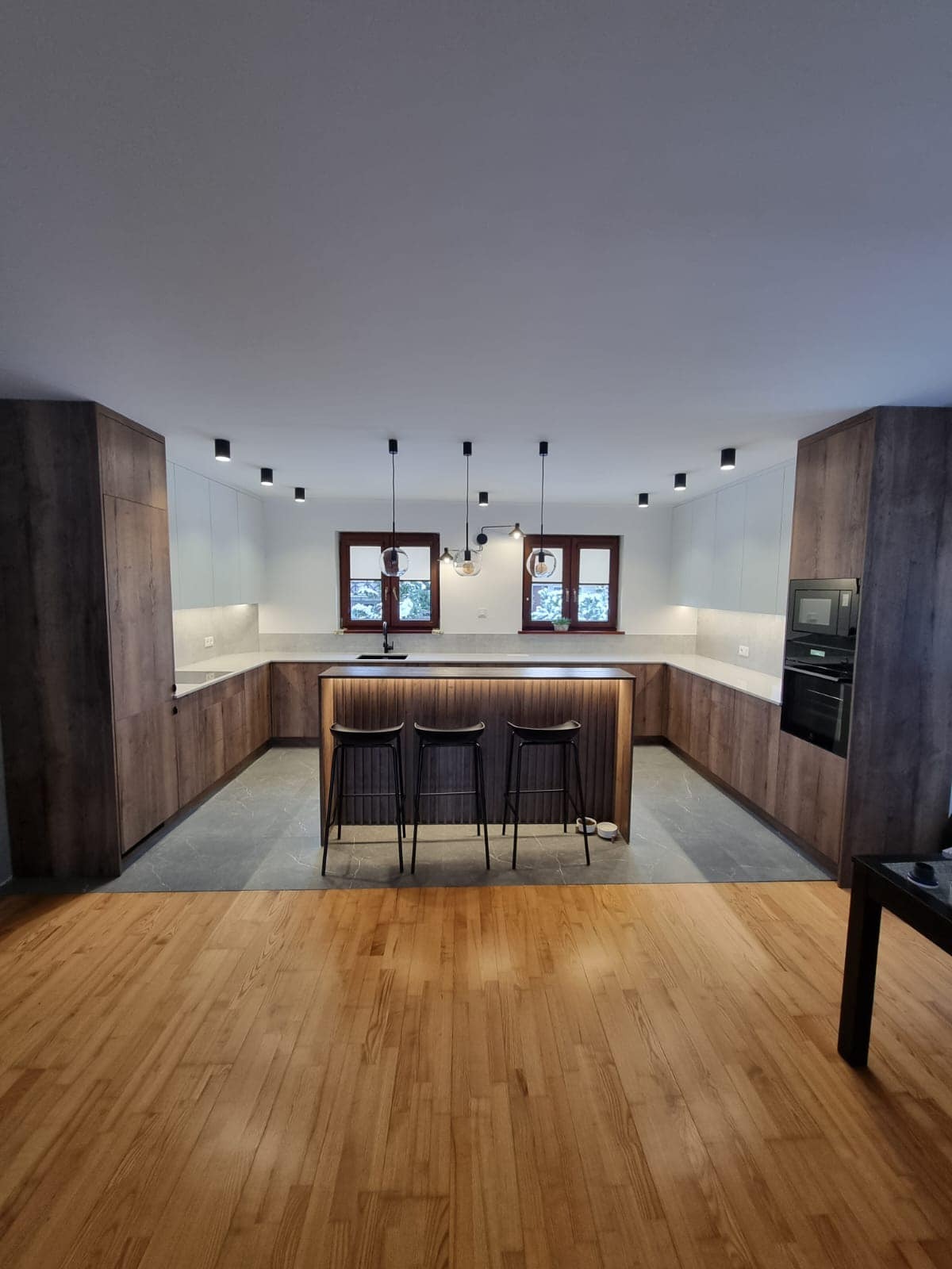 Nowoczesna kuchnia w kształcie litery U z wyspą i trzema hokerami, fronty szafek w odcieniach brązu z wyraźnym rysunkiem drewna, białe górne szafki, szara podłoga i drewniana podłoga w części...