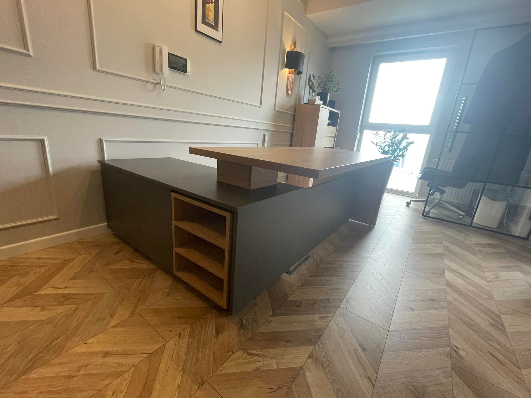 Nowoczesne biurko z ciemnoszarym korpusem i blatem z jasnego drewna, z otwartymi półkami, ustawione na parkiecie w jodełkę, w tle ściana z dekoracyjnymi listwami.