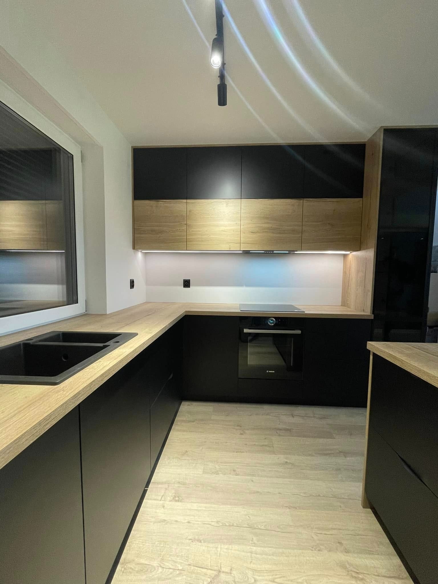 Nowoczesna kuchnia z czarnymi matowymi frontami i drewnianymi blatami, oświetlenie LED pod szafkami wiszącymi, czarny zlew podwieszany, widoczny fragment okna i podłogi w odcieniach drewna.