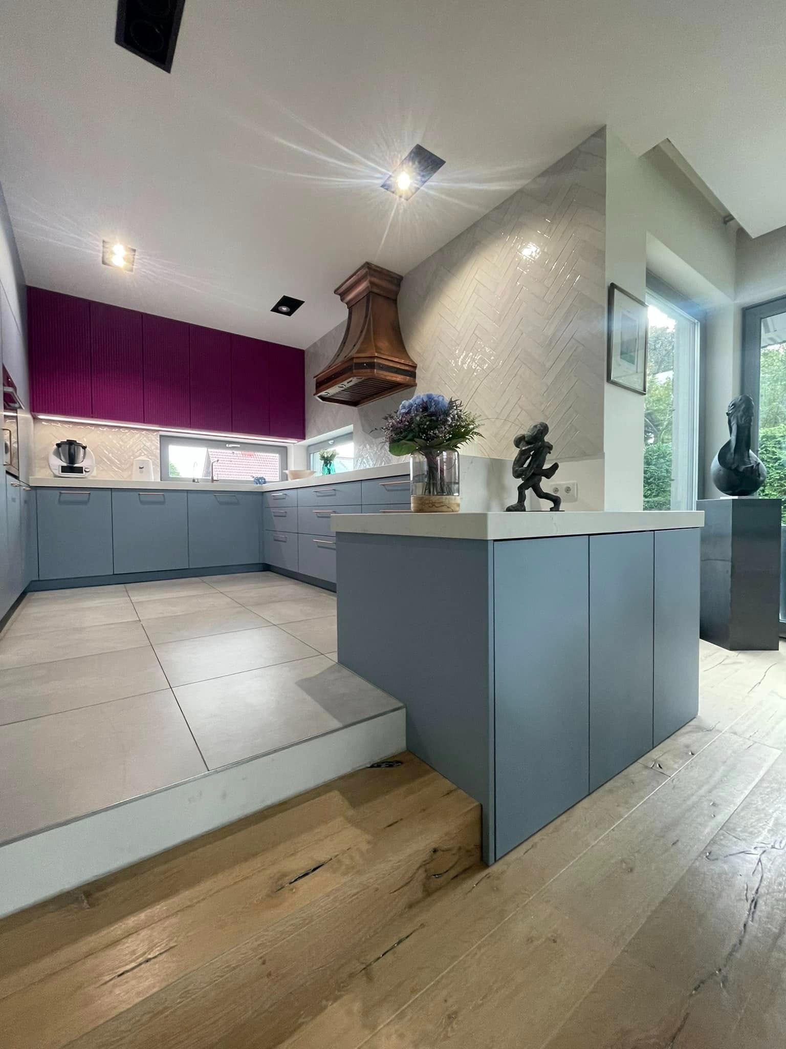 Nowoczesna kuchnia z szarymi szafkami, fioletowymi górnymi szafkami, drewnianym okapem i wyspą z dekoracjami, widoczna z poziomu podłogi z desek.