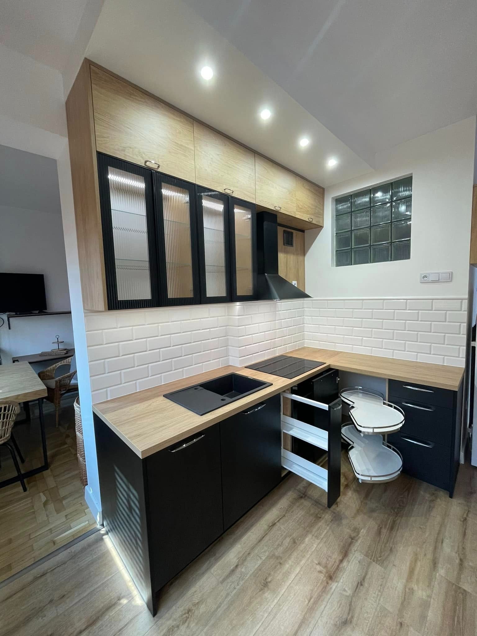 Nowoczesna kuchnia z drewnianymi blatami i czarnymi szafkami, widoczny otwarty narożny system przechowywania i szklane drzwi szafek wiszących z ryflowanym szkłem.