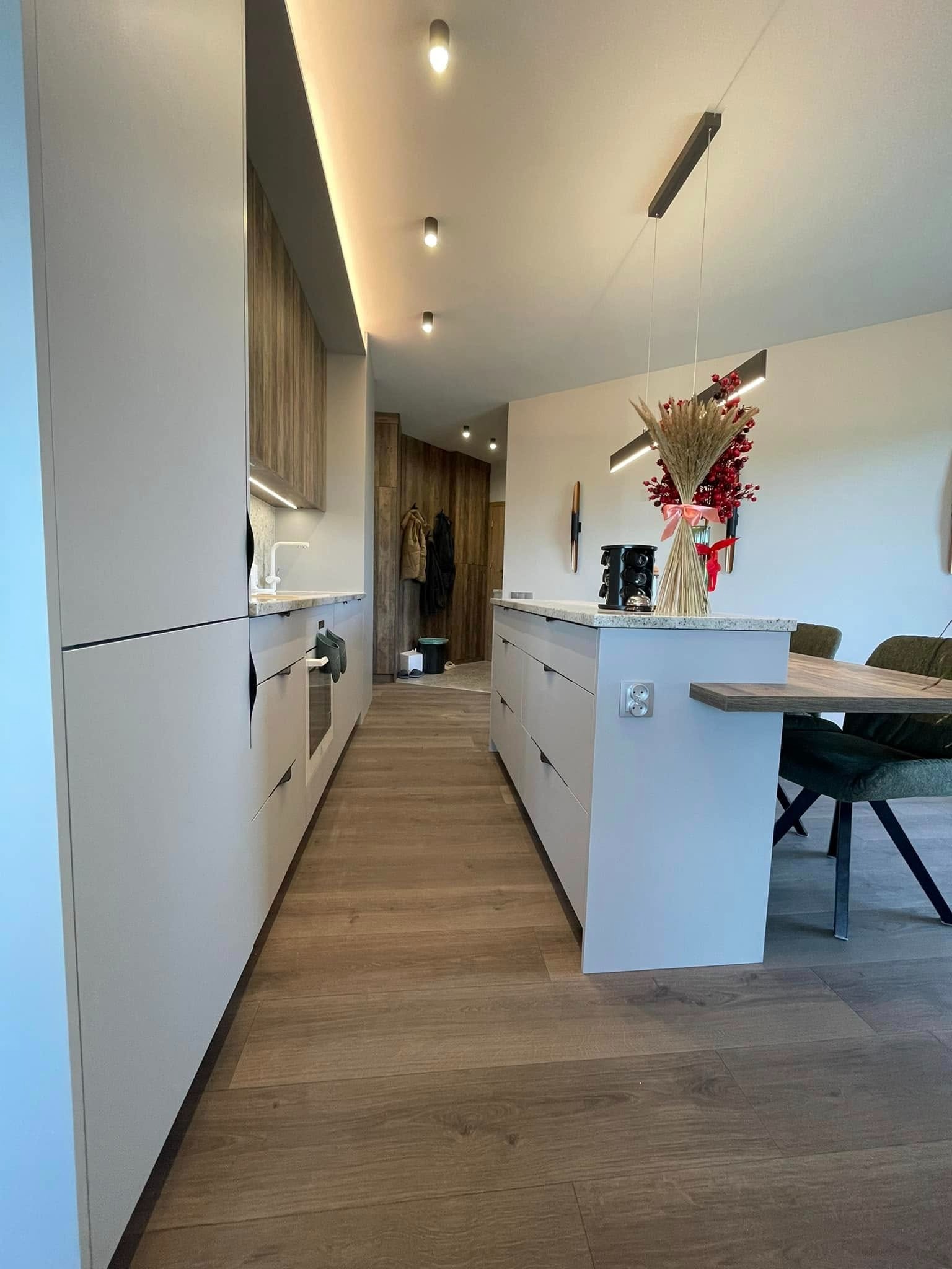 Nowoczesna kuchnia z szarymi frontami szafek i drewnianymi akcentami, oświetleniem LED podszafkowym i wyspą z blatem z jasnego kamienia, uzupełniona dekoracyjnym bukietem na wyspie i drewnianą...