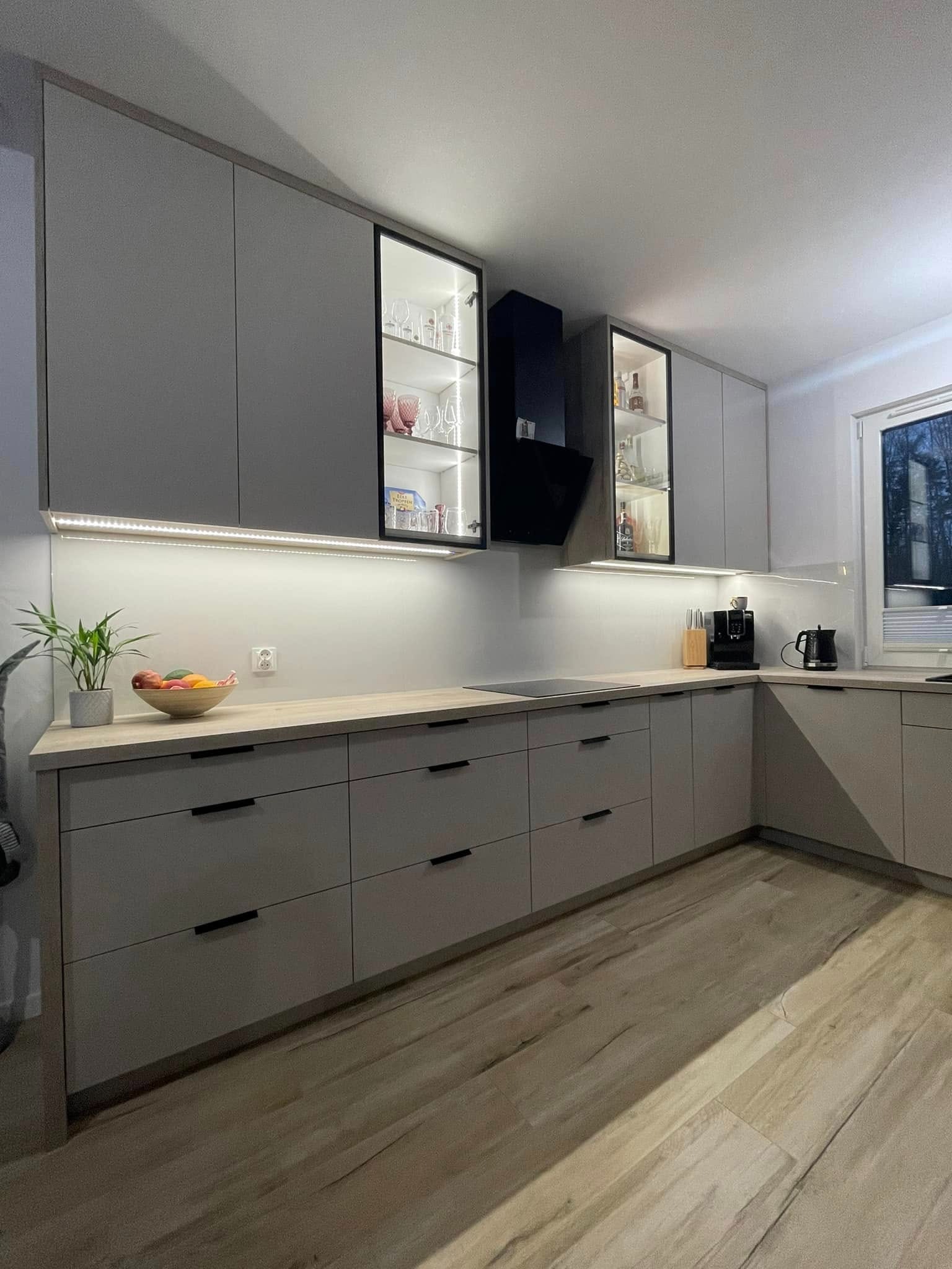 Nowoczesna kuchnia z szarymi szafkami, czarnymi uchwytami, przeszklonymi witrynami z podświetleniem LED, drewnianym blatem i drewnopodobną podłogą.