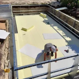 Budowa basenu: pracownik układa izolację termiczną i panele wewnątrz wykopu z metalową konstrukcją nośną, widoczne narzędzia i zapas materiałów.