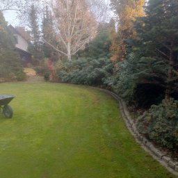 Ogrodnik - Zadbany trawnik z obrzeżem i taczką w ogrodzie. W tle drzewa i dom. Widok na równo skoszoną trawę i estetyczne wykończenie rabat.