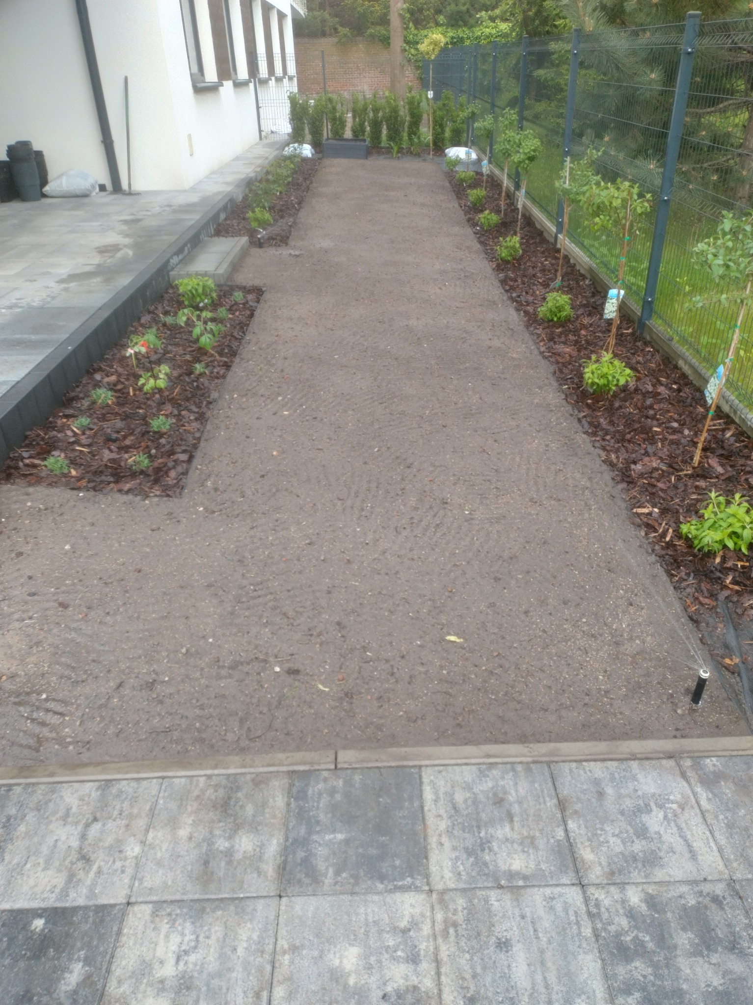 Ścieżka w ogrodzie z nowo posadzonymi drzewkami i krzewami, wyłożona korą. W tle zielona siatka ogrodzeniowa i budynek z białymi ścianami.