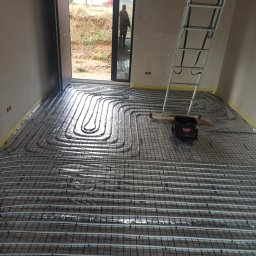 Usługi Hydrauliczne Radosław Szcześniak - Instalacja ogrzewania podłogowego: rury grzewcze ułożone na siatce w surowym wnętrzu, widoczna drabina i torba narzędziowa Makita, w tle otwarte drzwi z widokiem na teren budowy.