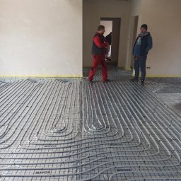 Usługi Hydrauliczne Radosław Szcześniak - Szare rury ogrzewania podłogowego ułożone na folii z nadrukiem Schilte, widoczni dwaj instalatorzy w tle, jeden w czerwonym kombinezonie, drugi w niebieskiej kurtce i czapce.