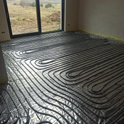 Usługi Hydrauliczne Radosław Szcześniak - Szare rury ogrzewania podłogowego ułożone na srebrnej folii z nadrukiem w nowym pomieszczeniu z dużym oknem wychodzącym na pole.